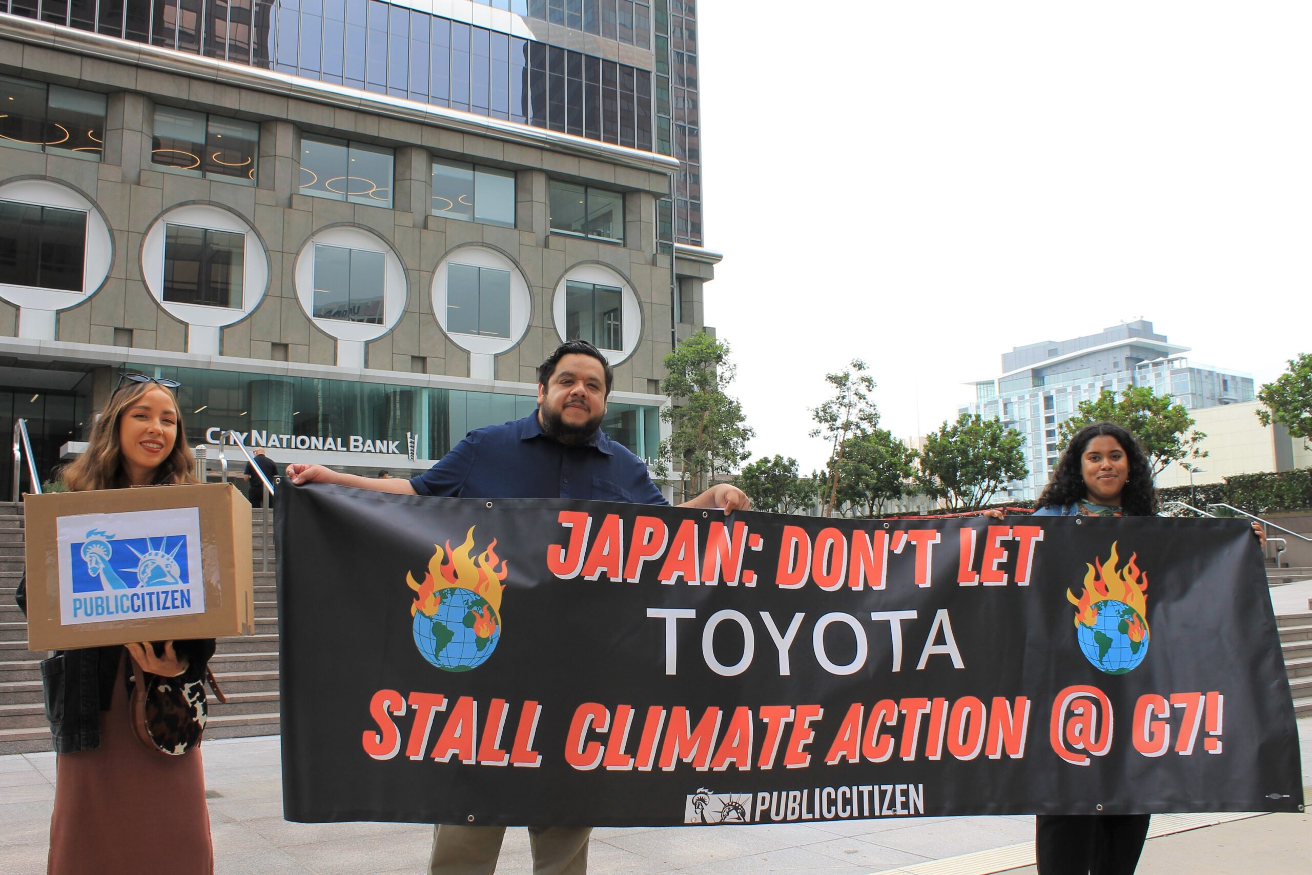 2 Public Citizen staff and 1 ally hold a banner saying, "Japan: don't let Toyota stall climate action @G7!"